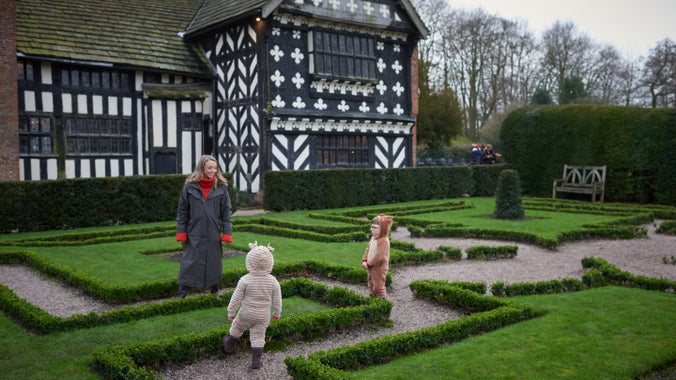 Two children wearing reindeer onesies and a woman stood in the formal garden outside of a black and white Tudor house.